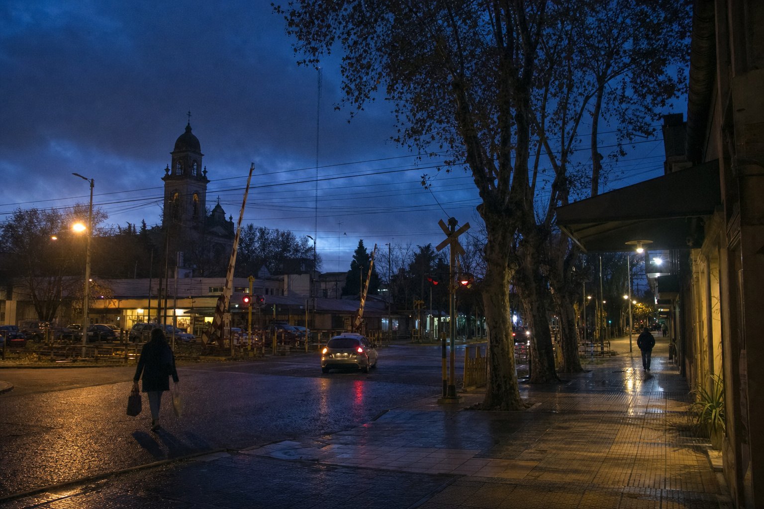 Calle céntrica de Morón al atardecer, con la torre de la iglesia y una barrera ferroviaria del Sarmiento.